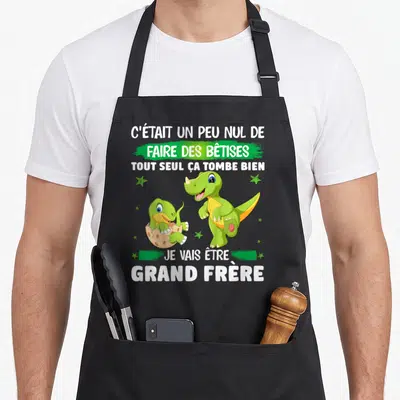 Tablier de cuisine noir avec deux dinosaures verts et l'annonce Je vais être grand frère imprimée en blanc.