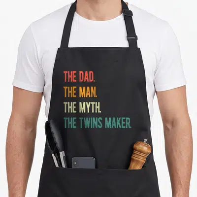 Tablier de cuisine noir avec le texte The Dad The Man The Myth The Twins Maker en lettres colorées verticales.