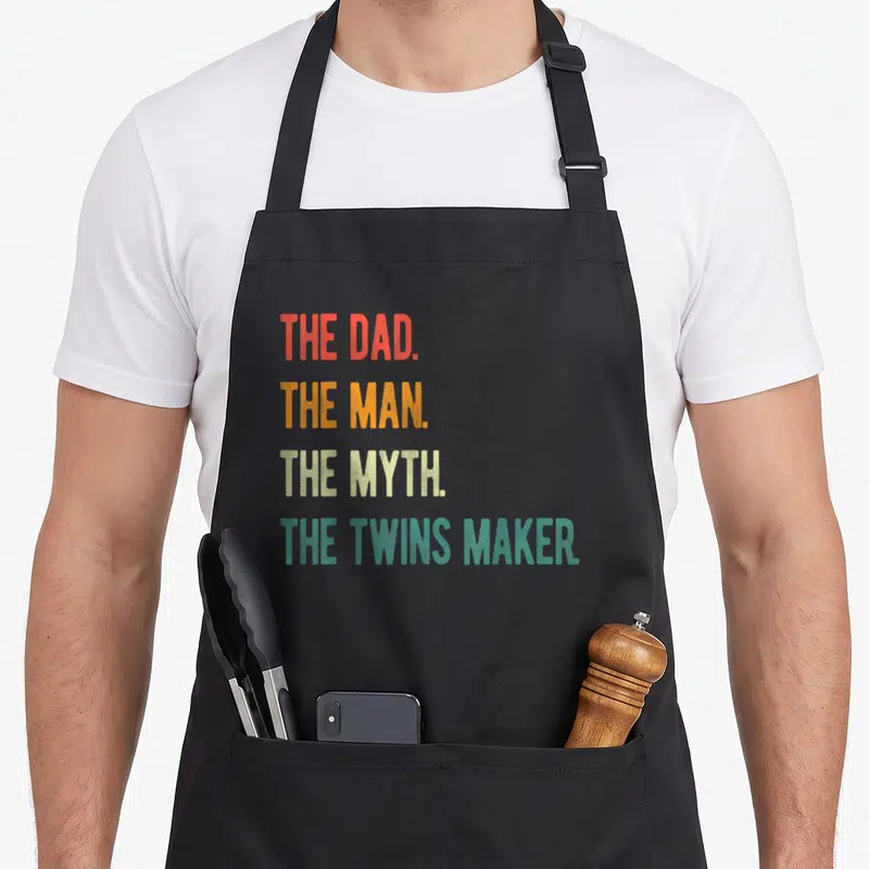 Tablier de cuisine noir avec le texte The Dad The Man The Myth The Twins Maker en lettres colorées verticales.