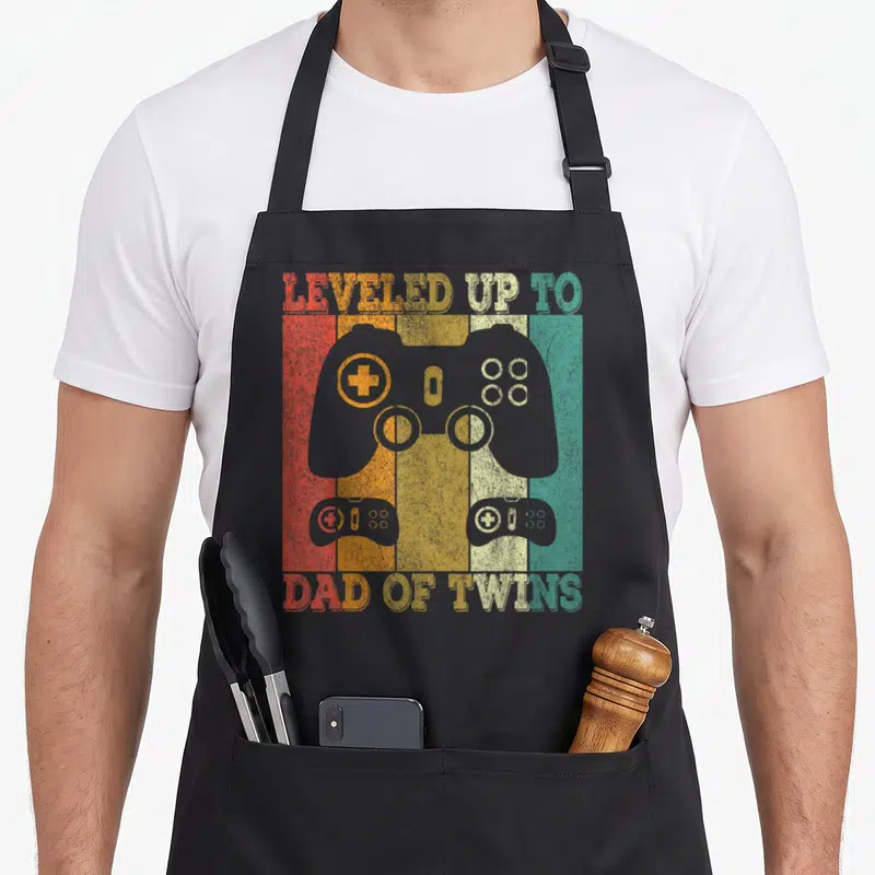 Tablier de cuisine noir avec illustration vintage de manette de jeu vidéo et texte en anglais coloré.