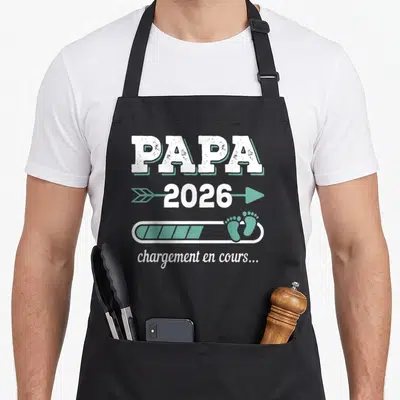 Tablier de cuisine noir avec l'inscription Papa 2026 chargement en cours et des petits pieds bleus.