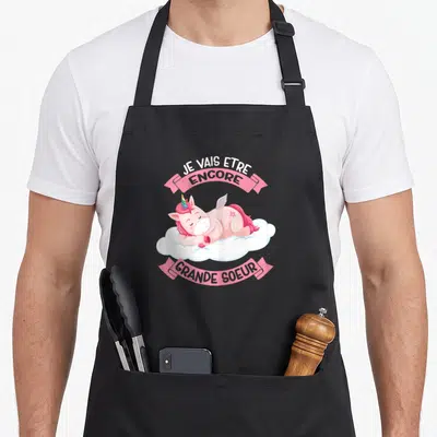 Tablier de cuisine noir avec une licorne rose dormant sur un nuage et l'annonce Je vais être encore grande sœur.
