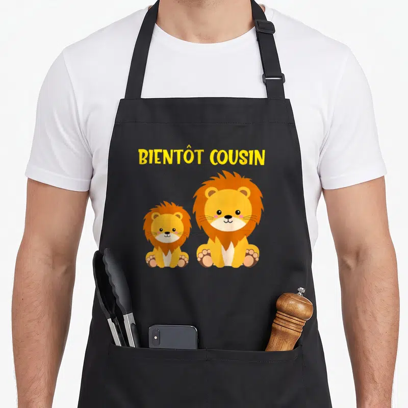 Tablier de cuisine noir avec deux lions orange et le texte Bientôt Cousin en jaune.