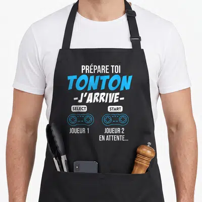 Tablier de cuisine noir avec l'inscription Prépare toi Tonton J'arrive et deux manettes de jeu vidéo bleues.