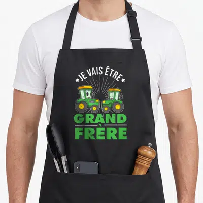 Tablier de cuisine noir avec deux tracteurs verts souriants et le texte Je vais être grand frère imprimé en blanc et vert.