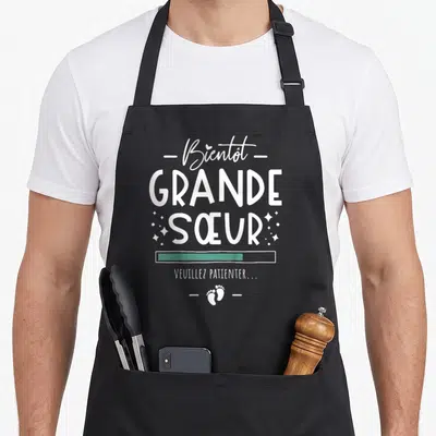Tablier de cuisine noir avec l'inscription Bientôt Grande Sœur Veuillez patienter et des empreintes de petits pieds en bas.