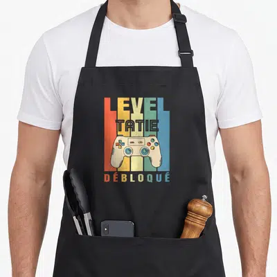 Tablier de cuisine noir avec illustration manette de jeu vidéo et texte Level Tatie Débloqué sur fond de bandes colorées verticales.