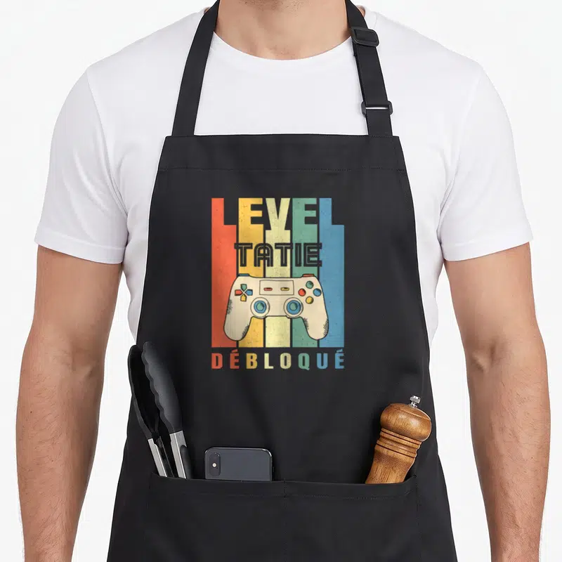 Tablier de cuisine noir avec illustration manette de jeu vidéo et texte Level Tatie Débloqué sur fond de bandes colorées verticales.