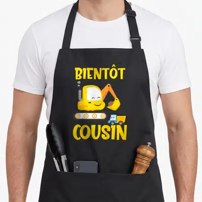 Tablier de cuisine noir avec une excavatrice jaune souriante et le texte Bientôt Cousin imprimé en lettres jaunes.