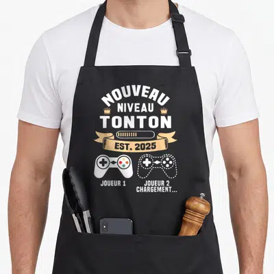 Tablier de cuisine noir avec inscription Nouveau Niveau Tonton Est 2025 et deux manettes de jeu vidéo stylisées.