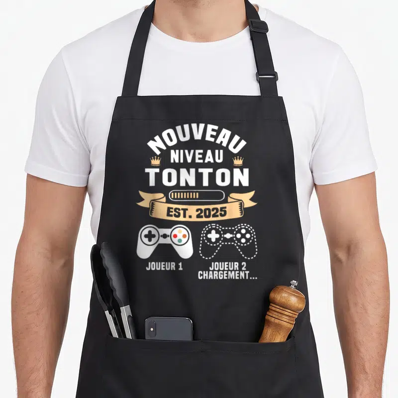 Tablier de cuisine noir avec inscription Nouveau Niveau Tonton Est 2025 et deux manettes de jeu vidéo stylisées.