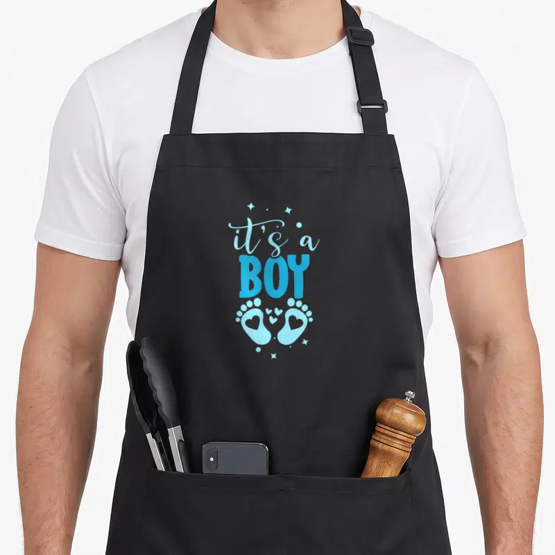 Tablier de cuisine noir avec le texte It's a Boy et deux petites empreintes de pieds bleues ornées de cœurs.