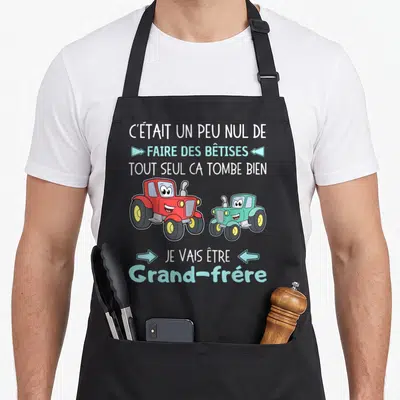 Tablier de cuisine noir avec deux tractors souriants et le message Je vais être grand-frère imprimé en bleu clair.