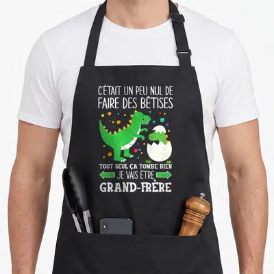 Tablier de cuisine noir avec un dinosaure vert et le texte Je vais être grand-frère entouré de pois colorés.