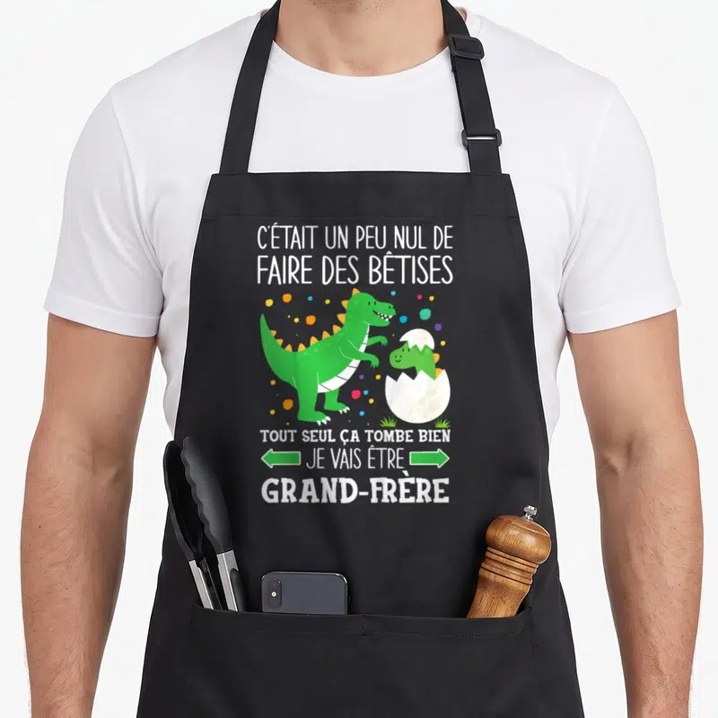 Tablier de cuisine noir avec un dinosaure vert et le texte Je vais être grand-frère entouré de pois colorés.
