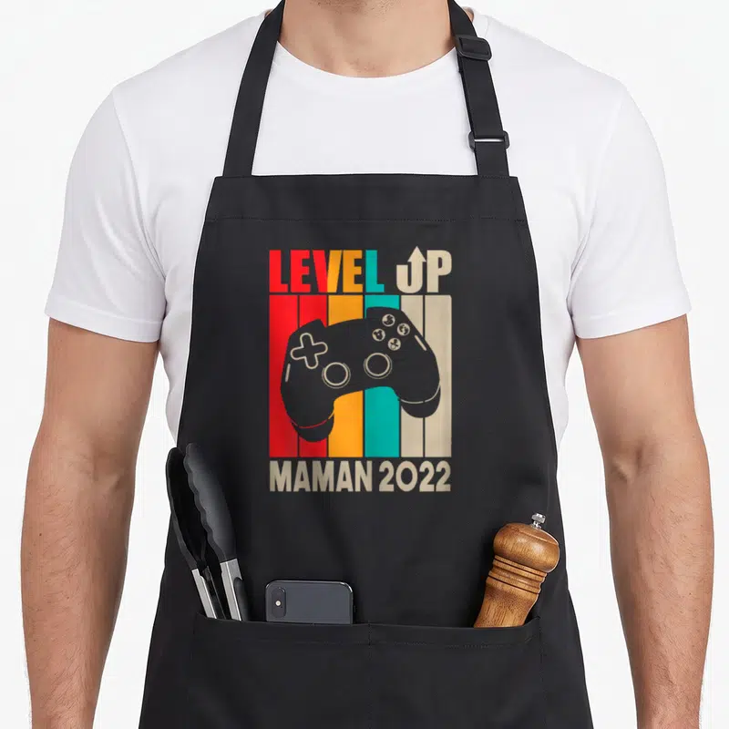 Tablier de cuisine noir avec illustration manette de jeu vidéo et texte Level Up Maman 2022 sur fond de bandes colorées.