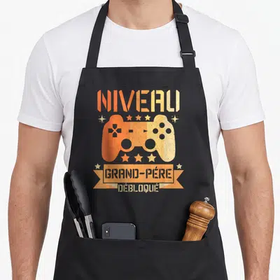 Tablier de cuisine noir avec manette de jeu orange et texte Niveau Grand-Père Débloqué imprimé au centre.
