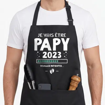Tablier de cuisine noir avec le texte blanc Je vais être papy 2023, une barre de progression verte et des petits pieds de bébé en bas.