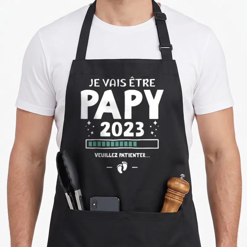 Tablier de cuisine noir avec le texte blanc Je vais être papy 2023, une barre de progression verte et des petits pieds de bébé en bas.
