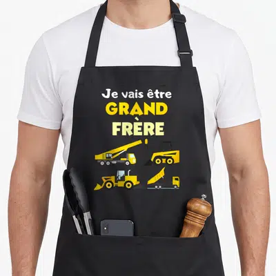 Tablier de cuisine noir avec « Je vais être grand frère » et quatre engins de chantier jaunes sur fond uni.