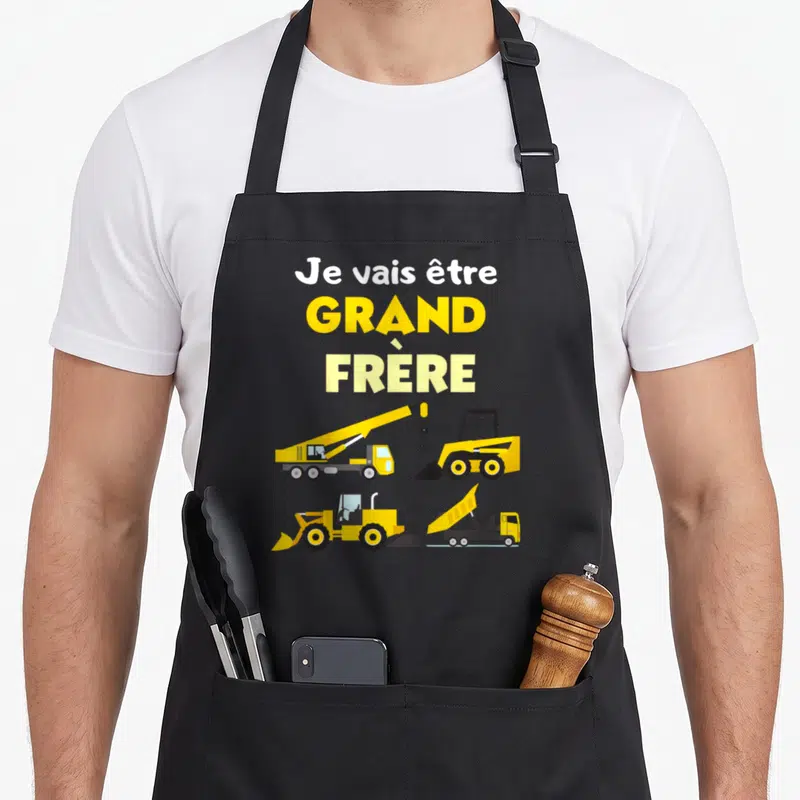 Tablier de cuisine noir avec « Je vais être grand frère » et quatre engins de chantier jaunes sur fond uni.