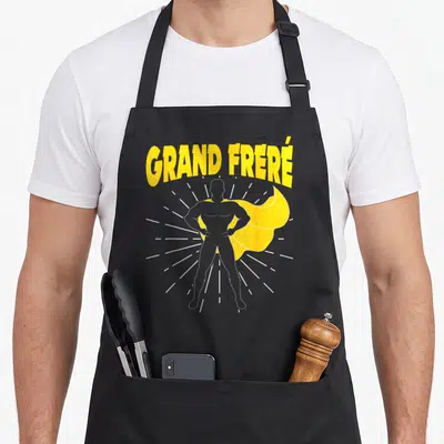Tablier de cuisine noir avec une illustration de super héros musclé et le texte Grand Fréré en lettres jaunes.