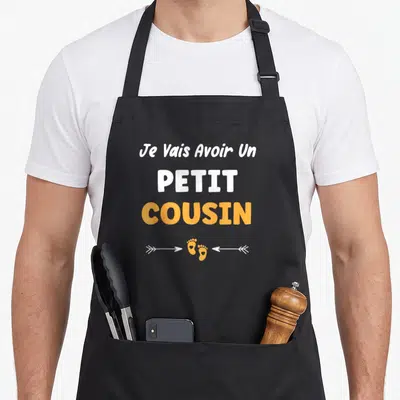 Tablier de cuisine noir avec le texte Je Vais Avoir Un PETIT COUSIN en blanc et orange surmontant des empreintes de pieds.