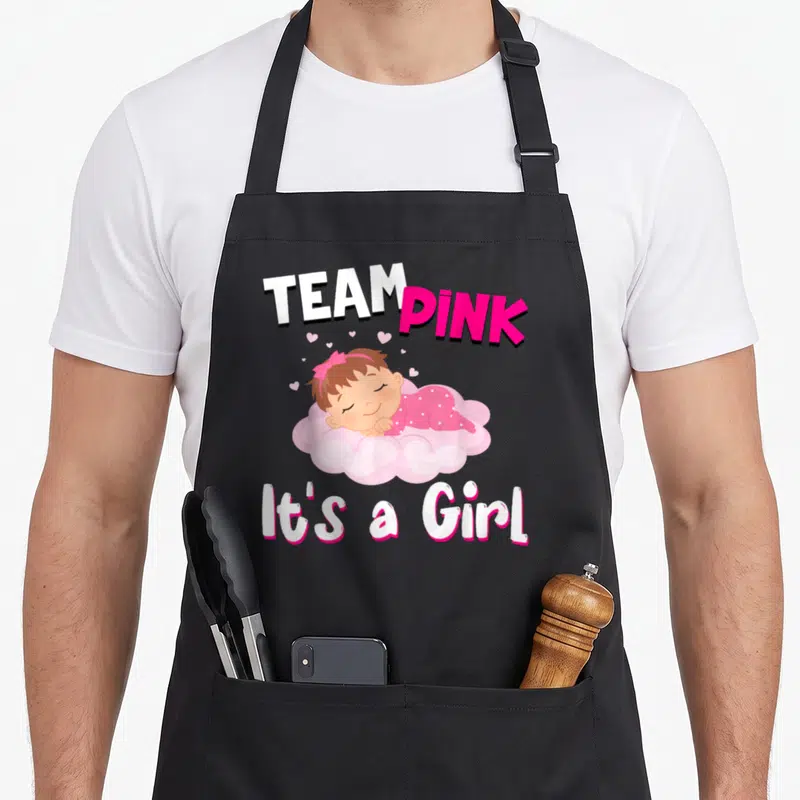 Tablier de cuisine noir avec illustration d'une bébé rose et les mots Team Pink It's a Girl imprimés en blanc et rose vif.