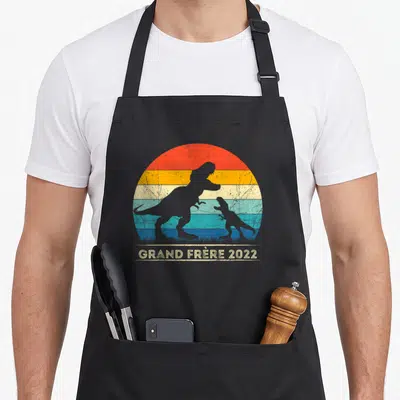 Tablier de cuisine noir avec silhouette de dinosaures et texte Grand Frère 2022 sur fond coloré vintage.