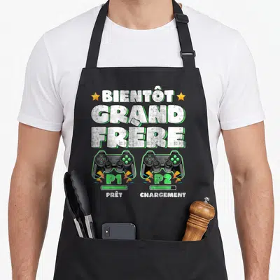 Tablier de cuisine noir avec texte Bientôt Grand Frère et deux manettes de jeu vidéo P1 Prêt P2 Chargement.