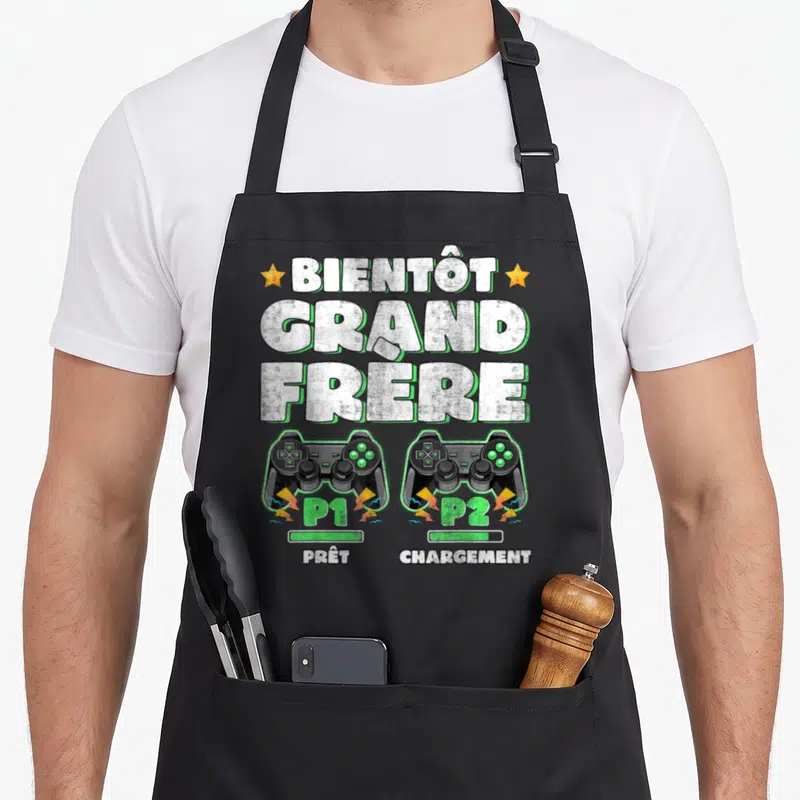 Tablier de cuisine noir avec texte Bientôt Grand Frère et deux manettes de jeu vidéo P1 Prêt P2 Chargement.