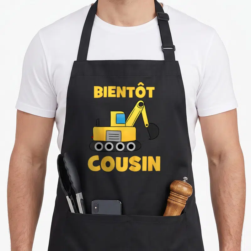 Tablier de cuisine noir avec illustration excavatrice jaune et texte Bientôt Cousin en lettres capitales jaunes.