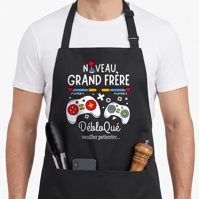 Tablier de cuisine noir avec deux manettes de jeu vidéo et texte Nouveau Grand Frère Débloqué.