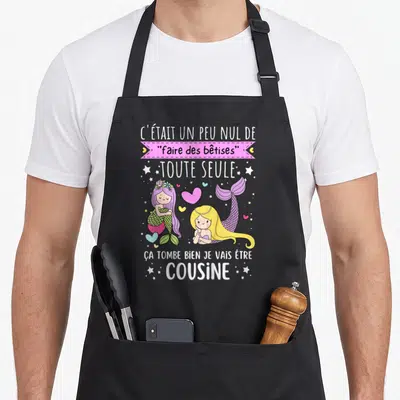 Tablier de cuisine noir avec deux sirènes colorées et le texte Je vais être cousine imprimé en blanc et rose.