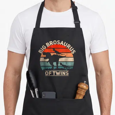 Tablier de cuisine noir avec silhouette dinosaure et texte Big Brosaurus of Twins sur fond de soleil rétro.