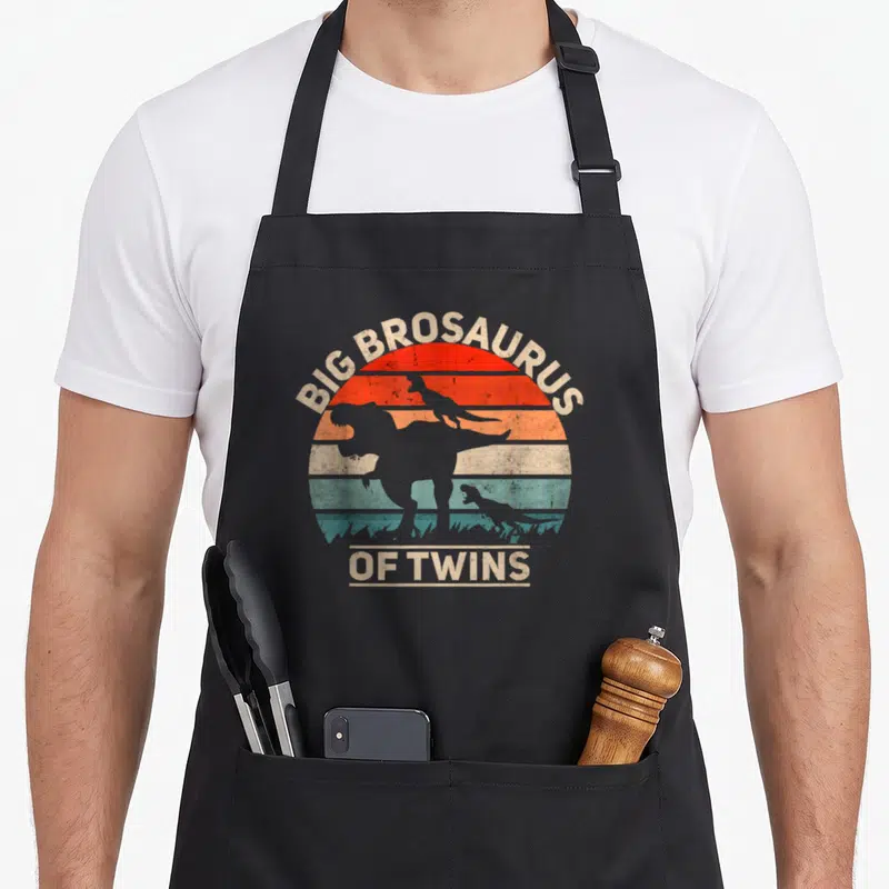 Tablier de cuisine noir avec silhouette dinosaure et texte Big Brosaurus of Twins sur fond de soleil rétro.