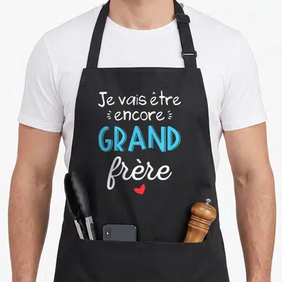 Tablier de cuisine noir avec le texte Je vais être encore grand frère en bleu et blanc surmonté d'un petit cœur rouge.
