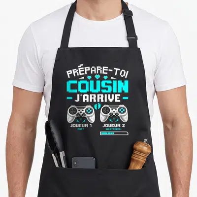 Tablier de cuisine noir avec l'inscription Prépare-toi Cousin J'arrive et deux manettes de jeu vidéo.