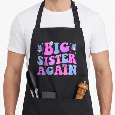 Tablier de cuisine noir avec inscription Big Sister Again en rose et bleu, orné de papillons et de fleurs stylisées.