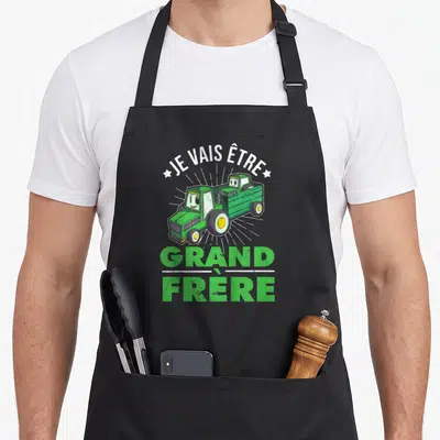Tablier de cuisine noir avec un tracteur vert et l'inscription Je vais être grand frère en lettres blanches et vertes.