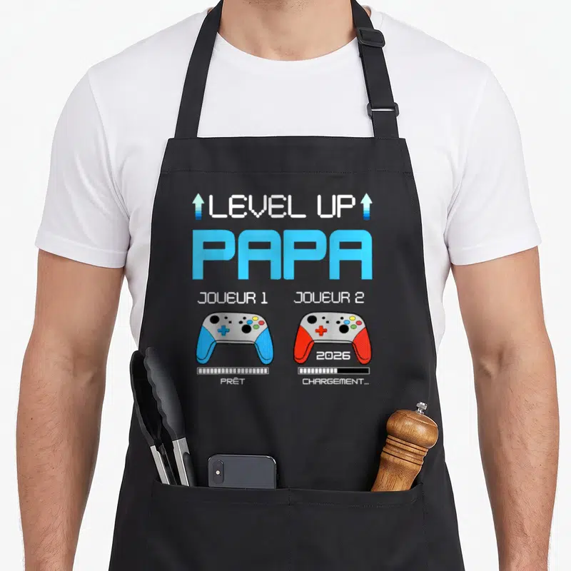 Tablier de cuisine noir avec illustration Level Up Papa 2026 et deux manettes de jeu vidéo colorées.