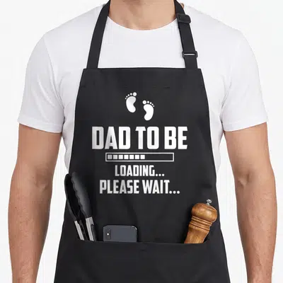 Tablier de cuisine noir avec texte blanc Dad To Be Loading Please Wait et deux petites empreintes de pieds au-dessus du message central.