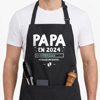 Tablier de cuisine noir avec inscription Papa en 2024 Veuillez patienter et empreintes de pieds blanches.