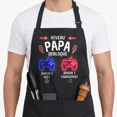 Tablier de cuisine noir avec texte Niveau Papa Débloqué et deux manettes de jeu vidéo colorées.