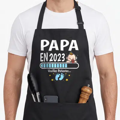 Tablier de cuisine noir avec texte Papa en cours 2023, barre de chargement bleue et petits pieds bleus entourés d'étoiles jaunes.