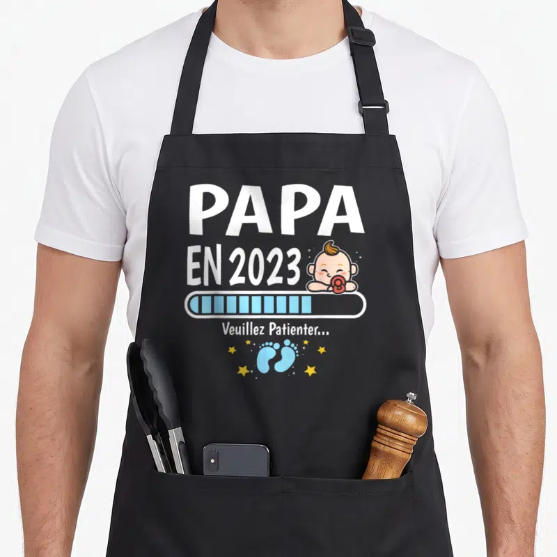 Tablier de cuisine noir avec texte Papa en cours 2023, barre de chargement bleue et petits pieds bleus entourés d'étoiles jaunes.