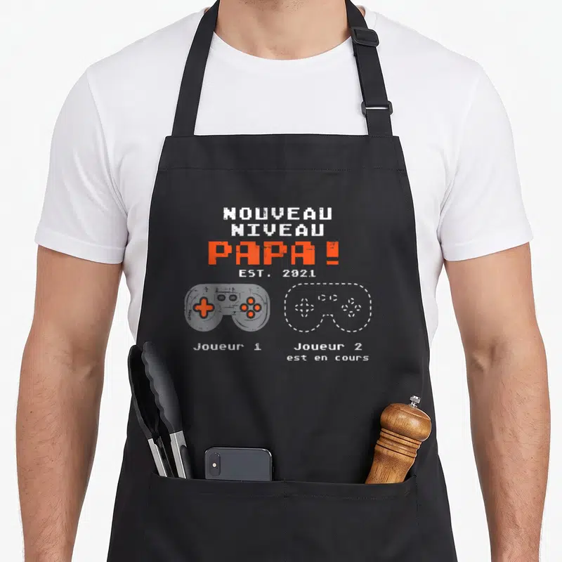 Tablier de cuisine noir avec inscription Nouveau Niveau Papa Est 2021 et deux manettes de jeu vidéo Jouleur 1 et Jouleur 2 est en cours.