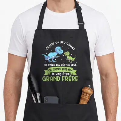 Tablier de cuisine noir avec deux dinosaures et le texte Je vais être grand frère imprimé en vert et blanc.