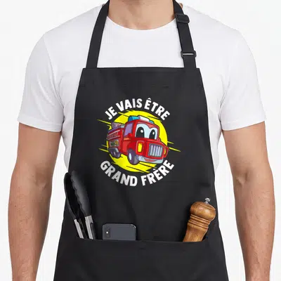 Tablier de cuisine noir avec illustration de camion de pompier rouge et texte blanc Je vais être grand frère sur fond jaune.