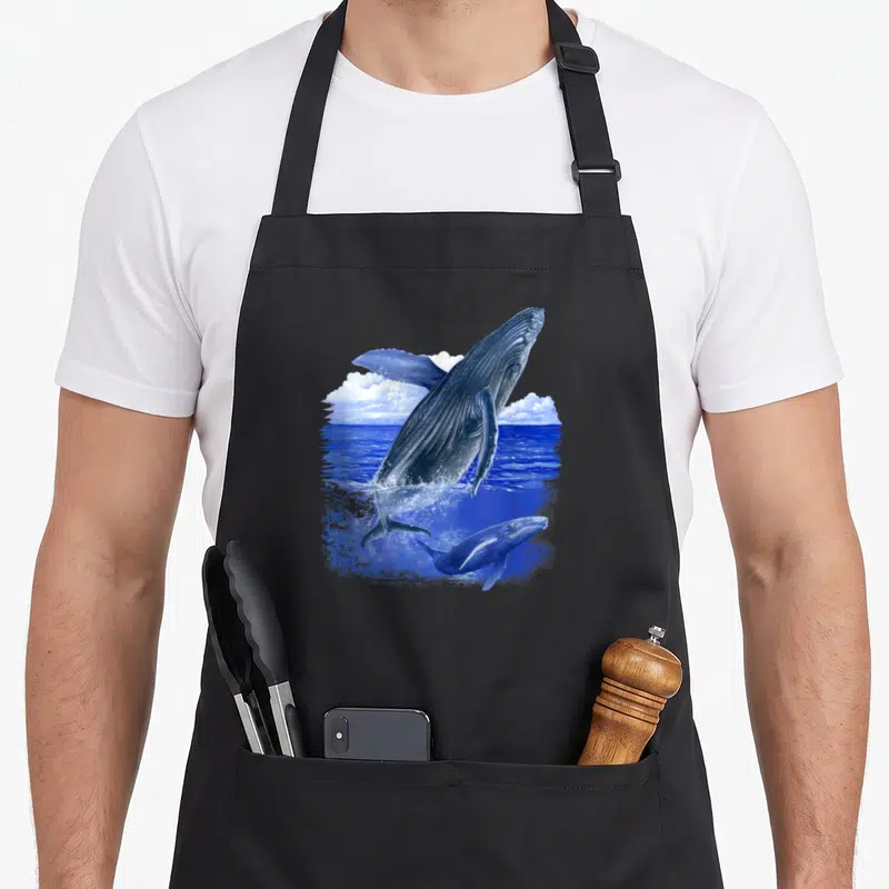 Tablier de cuisine noir avec une baleine et un bébé baleine sautant dans l'eau bleue sous des nuages blancs.
